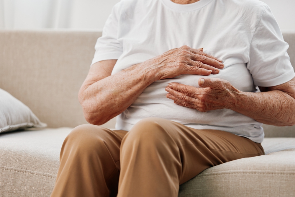 An elderly person sitting on a couch with a hand placed on their chest