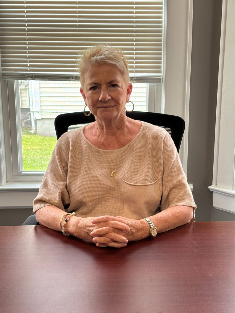 A headshot of Joy Thompson sitting at a table. Her hands are folded in front of her and there is a window open behind her.