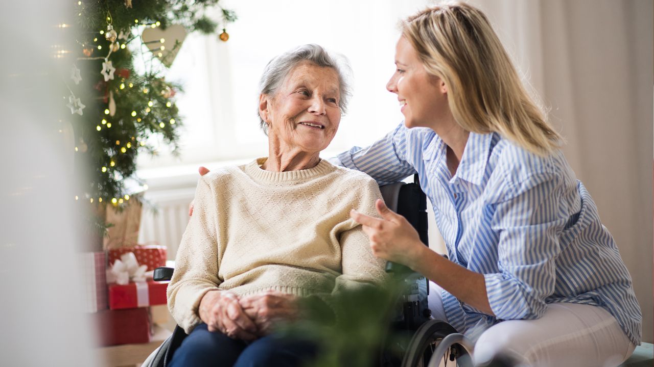 An elderly woman in a wheelchair smiles at a younger caregiver who is leaning towards her, both near a decorated Christmas tree with presents underneath. The scene highlights the warmth and joy brought by dedicated elder home care services during the festive season.