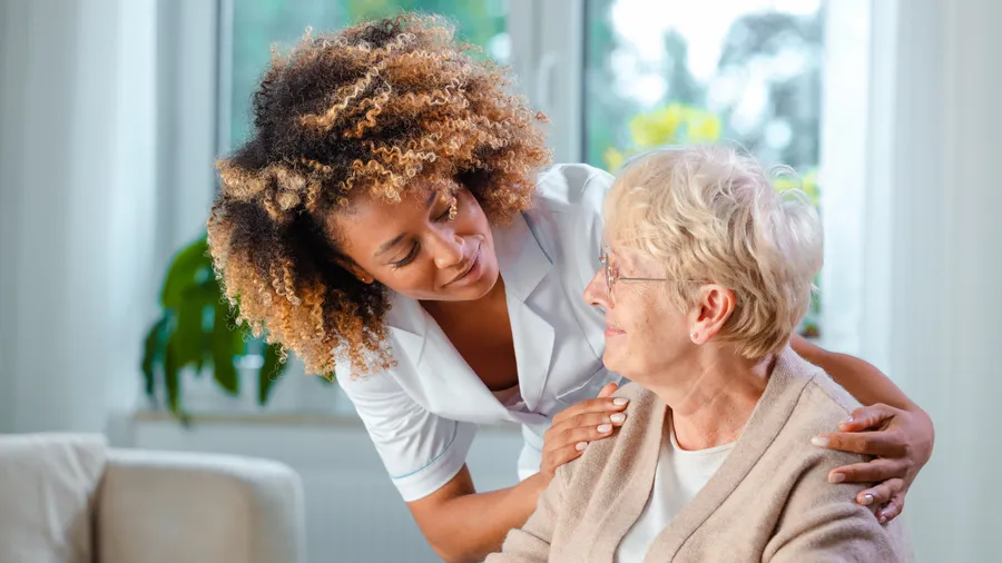 A nurse with curly hair gently holds the shoulders of an elderly woman with glasses, offering comfort. The warm interaction, a true example of home care, takes place indoors with a window view in the background.