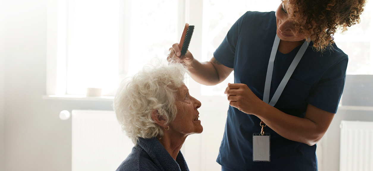 A caregiver in blue scrubs brushes the hair of an elderly person with white hair, who is seated in a well-lit room, showcasing the quality of elder home care services.