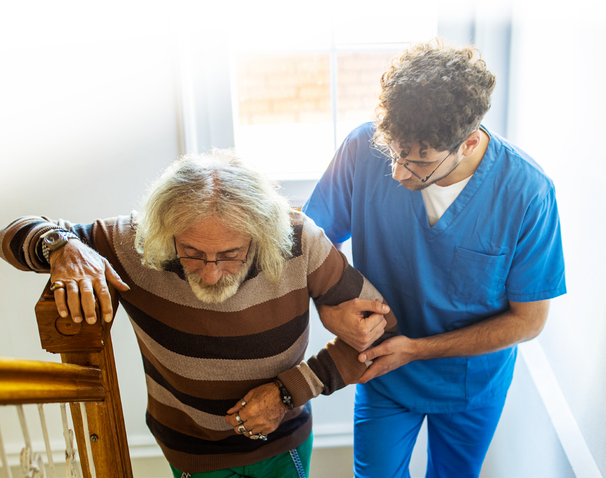 A healthcare worker in blue scrubs assists an elderly man with gray hair and a striped sweater up a staircase, showcasing the attentive support provided by elder home care services.