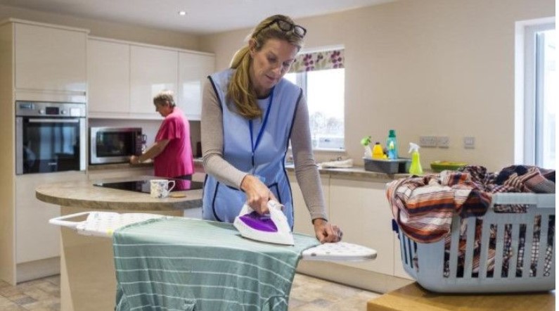 Two women in a kitchen: one is ironing clothes on an ironing board, and the other is using a microwave. A laundry basket is on the counter, showcasing the seamless integration of personal care services in daily life.