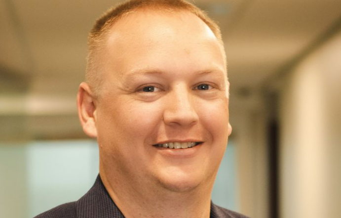 A man with short light hair, wearing a dark collared shirt, is smiling in an indoor setting with a blurred background, likely in a facility that offers home health care service.