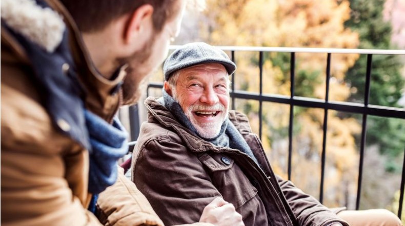 An elderly man in a brown jacket and cap smiles while sitting on a balcony, speaking with a younger person in a brown coat, exemplifying the warmth of home care.