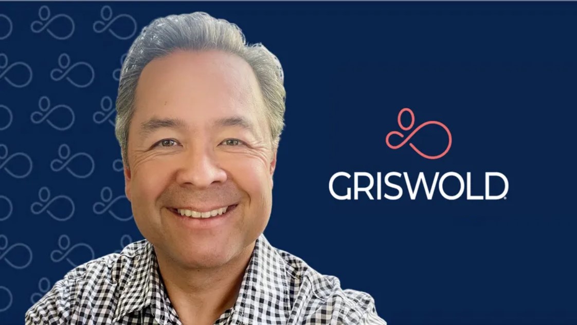 Man smiling in front of a blue background with the Griswold logo on it. He is wearing a checkered shirt, symbolizing his commitment to elder home care services.