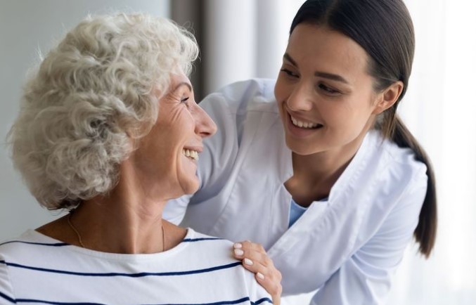 A smiling elderly woman with curly gray hair shares a cheerful moment with a caregiver from home care services in a white uniform, who has her hand on the woman's shoulder.