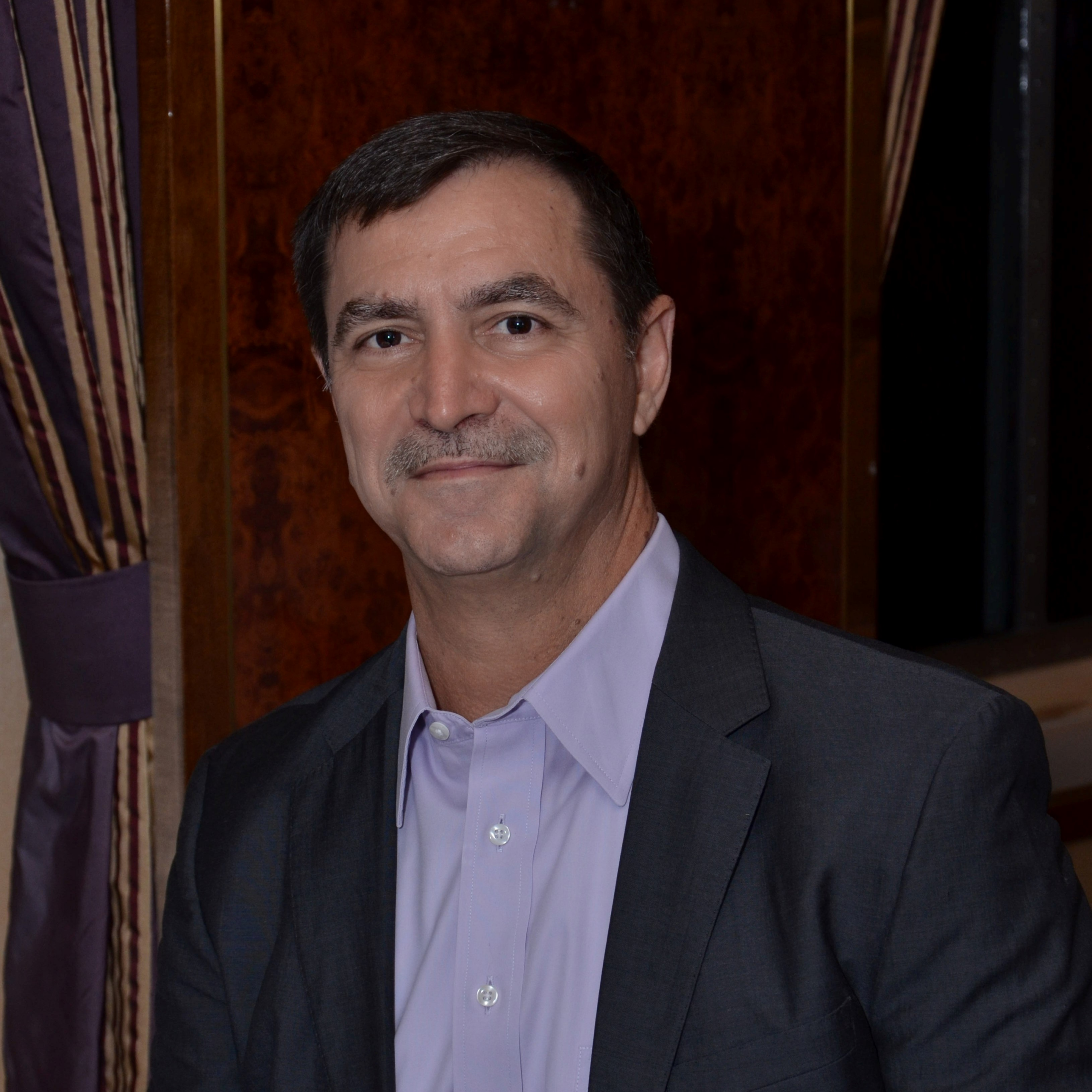 A man wearing a suit and light-colored shirt stands in front of a wood-paneled background with draped curtains visible on the side, embodying the professionalism and dedication one might expect from someone offering home health care services.