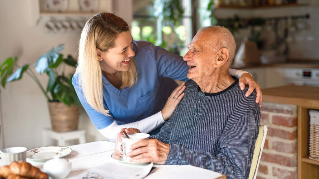 A nurse in blue scrubs, smiling and holding an elderly man's shoulder, while he sits at a table holding a cup in a well-lit kitchen—a beautiful moment of personal care services.