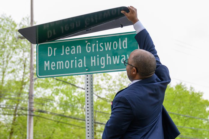 A man in a dark suit is unveiling a green road sign that reads "Dr Jean Griswold Memorial Highway" with trees and power lines visible in the background, honoring Dr. Griswold's contributions to elder home care services.