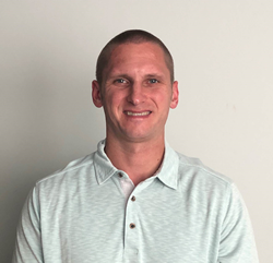 A person with short hair and a light blue collared shirt, perhaps representing personal care services, smiles at the camera against a plain background.