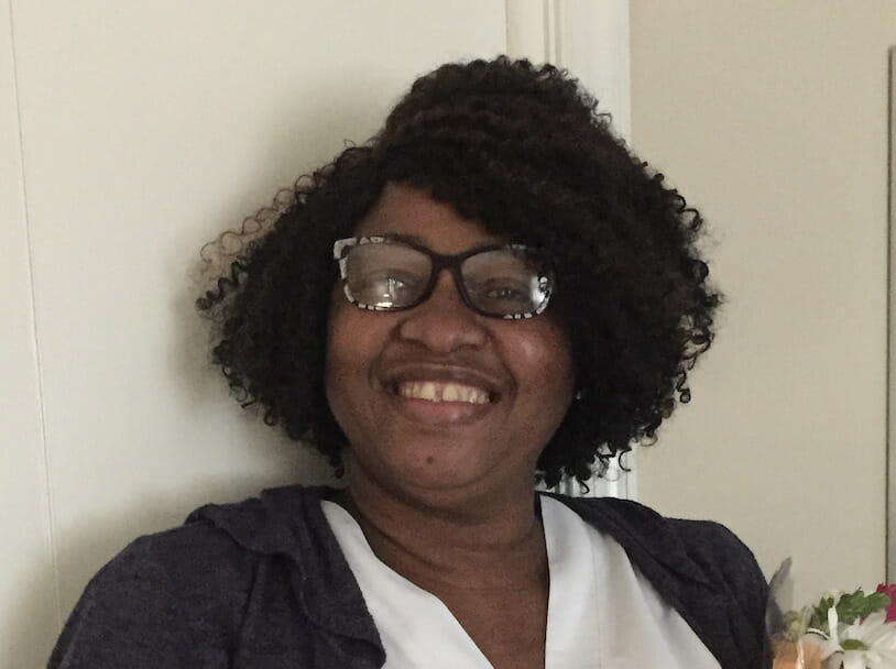 A person with curly hair and glasses is smiling. They are wearing a white top and a dark cardigan, embodying the warmth one might expect from someone involved in personal care services.