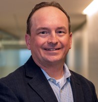 A man with short hair and a smile wears a dark blazer and light blue shirt, standing in a well-lit hallway, ready to discuss caregiver services.