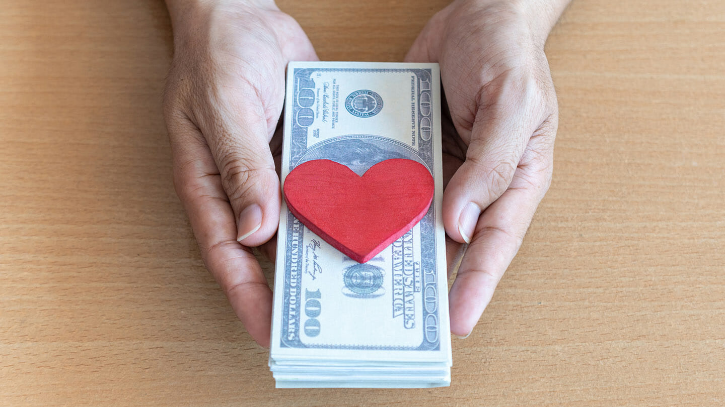 Hands holding a stack of 100-dollar bills with a red wooden heart placed on top, symbolizing the compassion and value found in quality home care services, over a wooden surface.