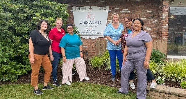 A group of six women standing and smiling in front of a brick building with a sign that reads "Criswold Home Care." They appear to be posing for a group photo, showcasing their dedication to providing exceptional elder home care services.