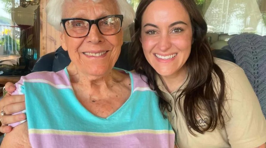 An older woman wearing glasses and a striped shirt sits next to a younger woman in a beige shirt; both are smiling at the camera, showcasing the warmth of care pairing.