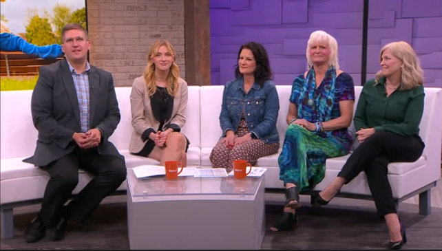 Five people are seated on a white couch in a TV studio, discussing the benefits of home health care services. Two orange mugs and papers are on a glass table in front of them. The background features a brick wall and a purple partition, adding to the engaging atmosphere.