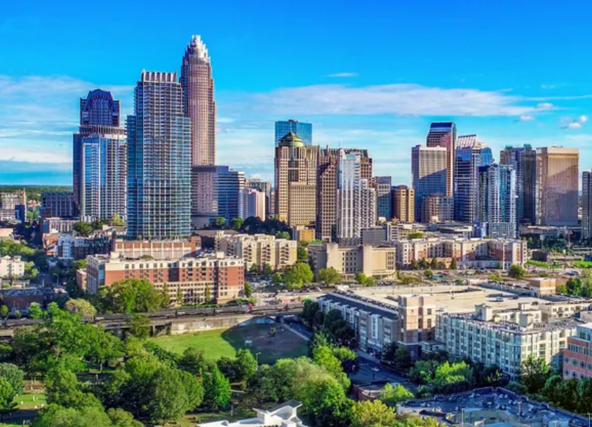 A city skyline with numerous tall buildings and skyscrapers is accompanied by a foreground of green spaces and residential areas, where home health care services ensure the well-being of residents under a clear blue sky.