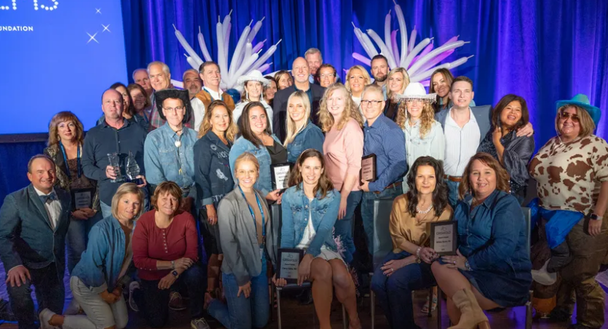 A group of people posing for a photo on a stage with blue curtains, some wearing headpieces. Several hold plaques celebrating their achievements in elder home care services. The backdrop displays a lit-up screen.