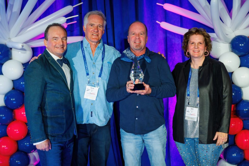 Four individuals stand together, smiling, with one person holding an award. They are in front of a backdrop featuring balloons and blue curtains, celebrating excellence in caregiver services.