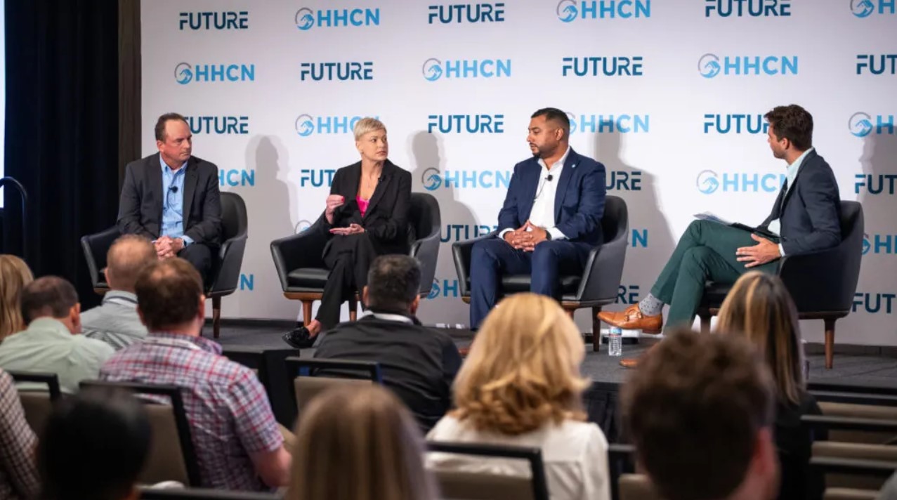 Four people are seated on a stage engaged in a panel discussion about elder home care services at a conference, with an audience in attendance.