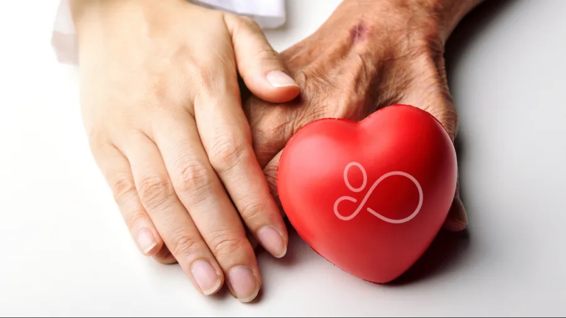 Two hands, one young and one elderly, rest on a surface together next to a red heart-shaped object with a decorative symbol, embodying the compassion and support central to home health care services.