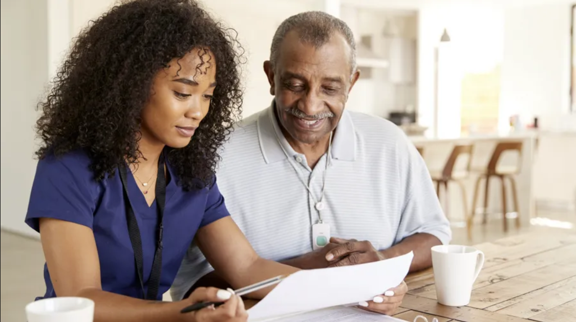 A woman in blue scrubs sits at a table, holding a pen and reviewing a document with an older man, who is smiling. Two mugs are on the table, illustrating the warmth of caregiver services.