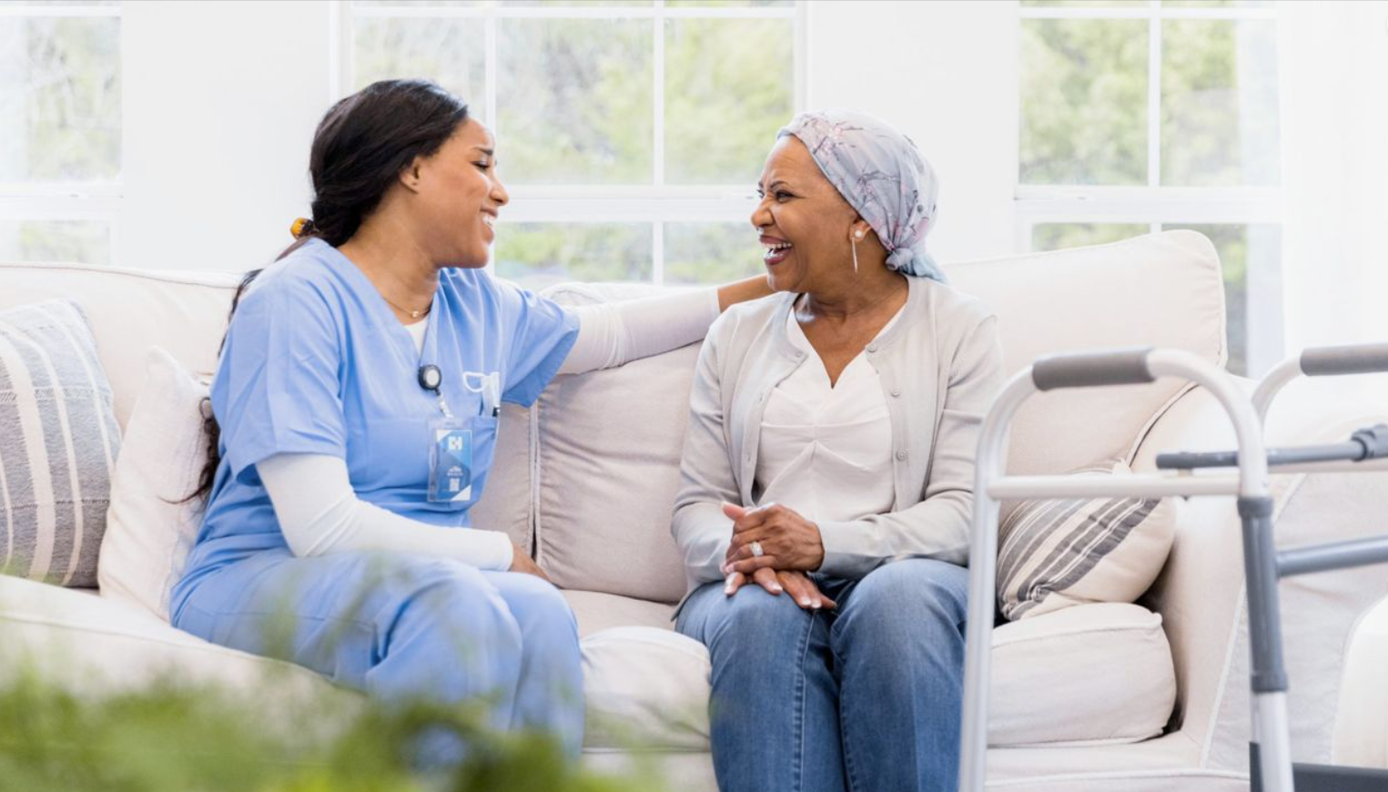 A healthcare worker in blue scrubs sits on a couch, smiling and touching the shoulder of an older woman with a headscarf, who holds a walking frame and smiles back, showcasing the warmth and compassion of elder home care services.