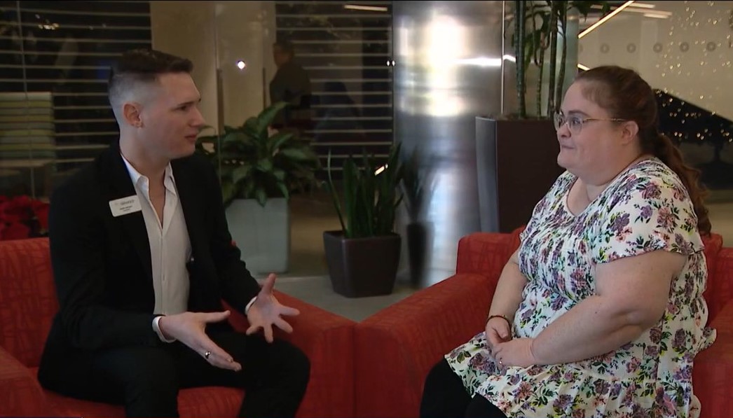 A man in a black suit and white shirt converses with a woman in a floral dress seated on a red couch in what appears to be a modern office lobby, possibly discussing elder home care services.