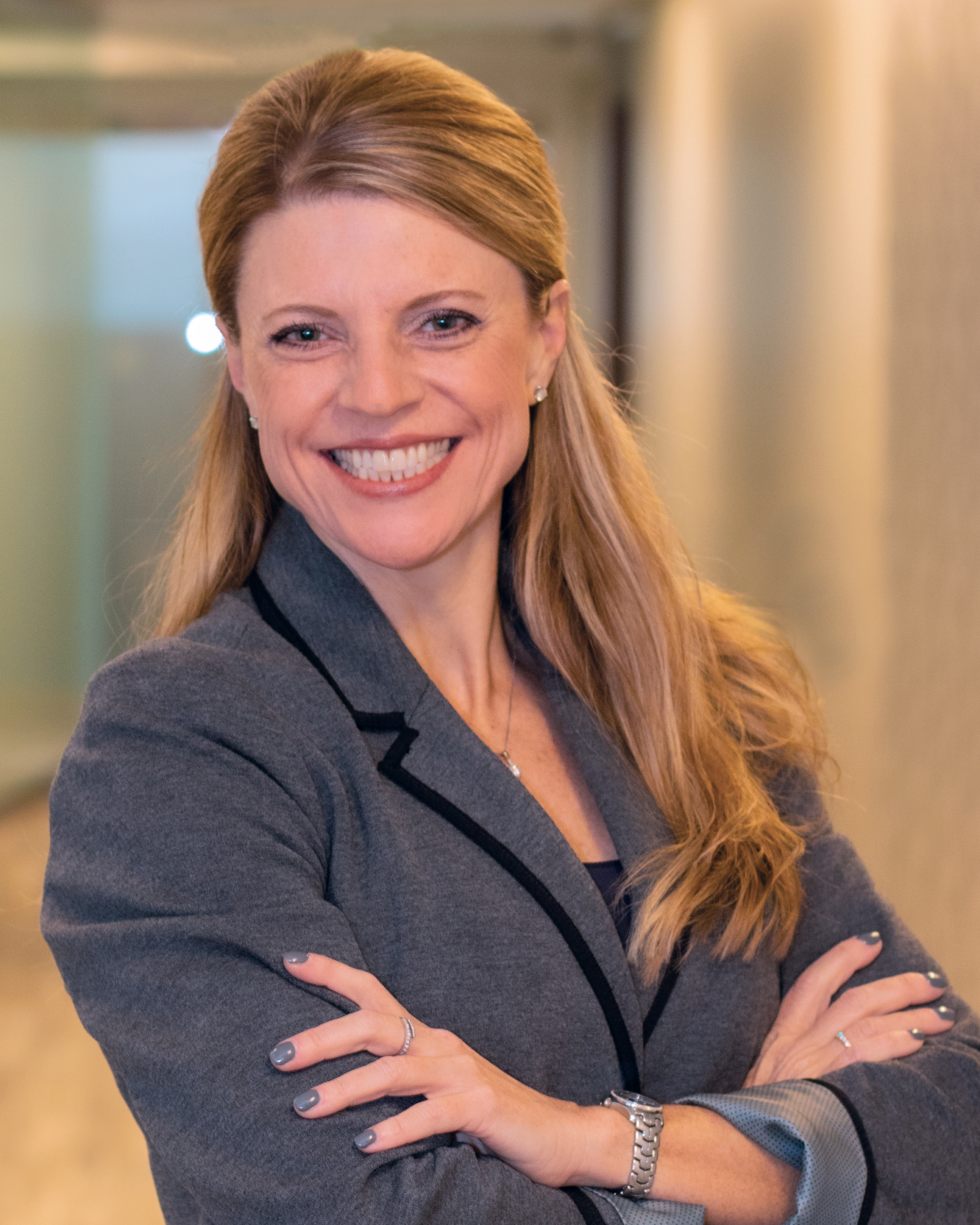 A smiling woman with long blonde hair and a gray blazer stands with arms crossed in a hallway, facing the camera, embodying professionalism and warmth often associated with elder home care services.