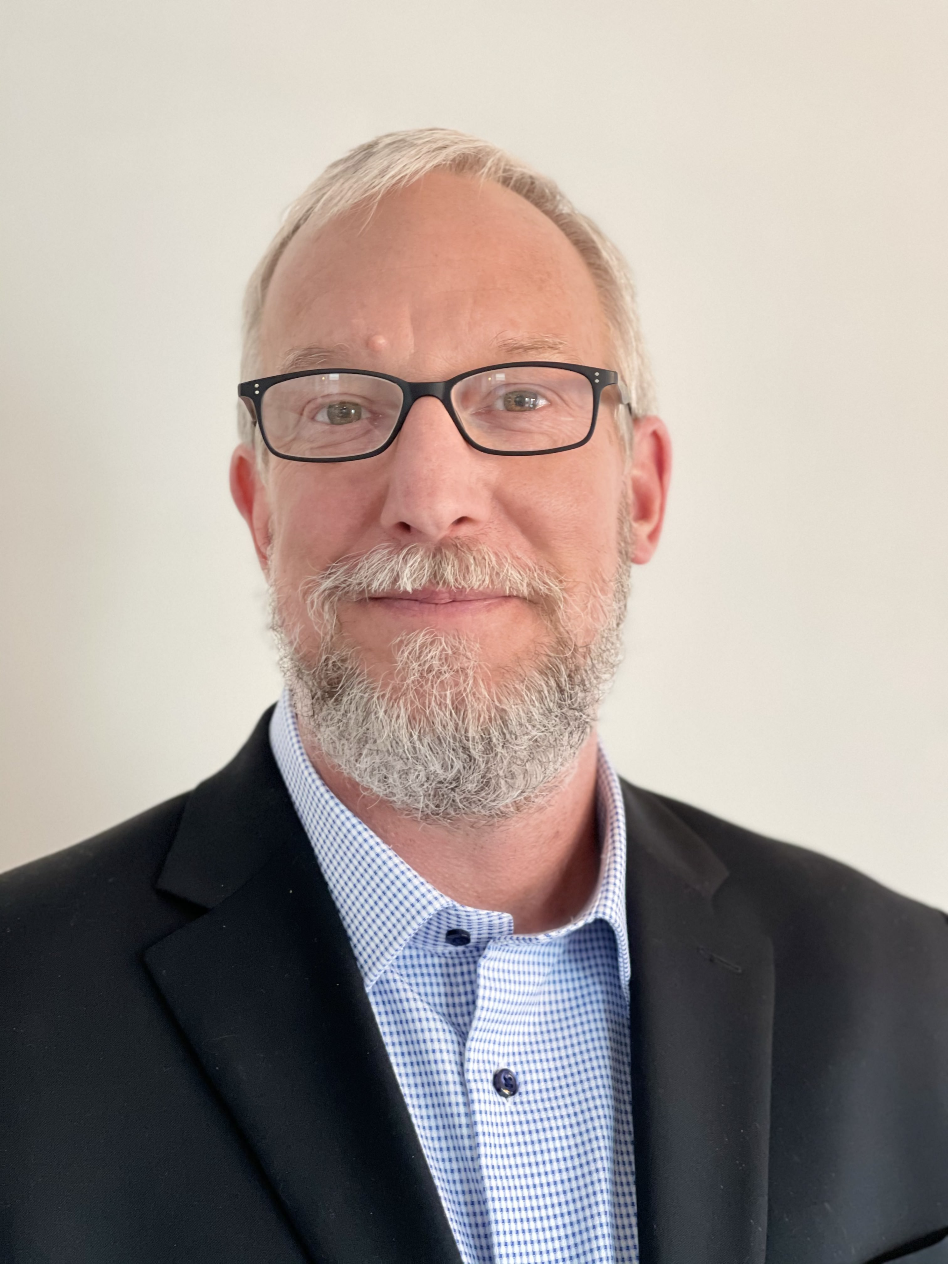 A man with glasses and a grey beard is wearing a dark suit jacket over a light blue, checked dress shirt. He is standing against a plain white background, looking poised and professional as if ready to discuss elder home care services.