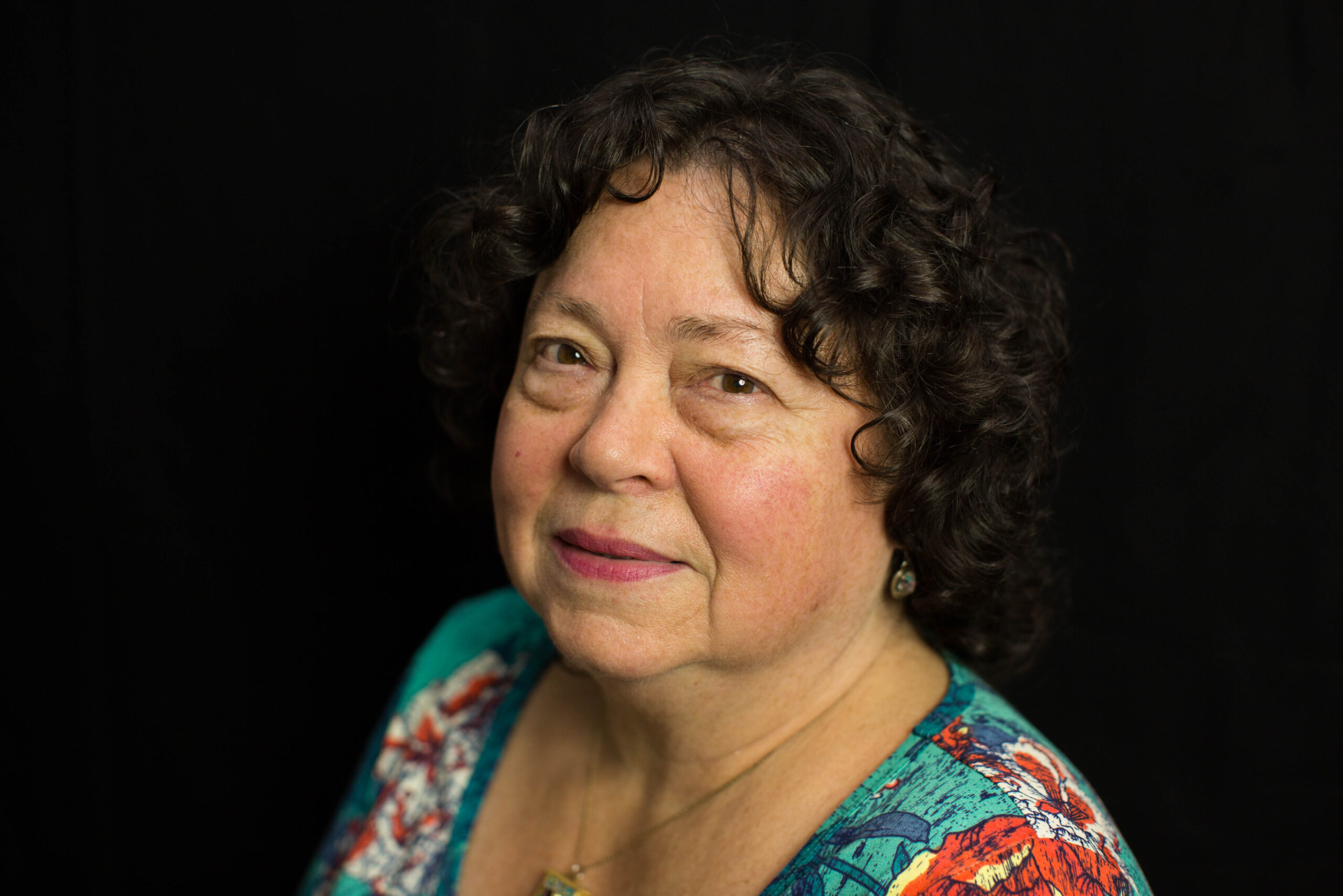 A woman with short curly hair and a patterned top poses for a portrait against a black background, embodying the comforting essence often found in dedicated home care services.