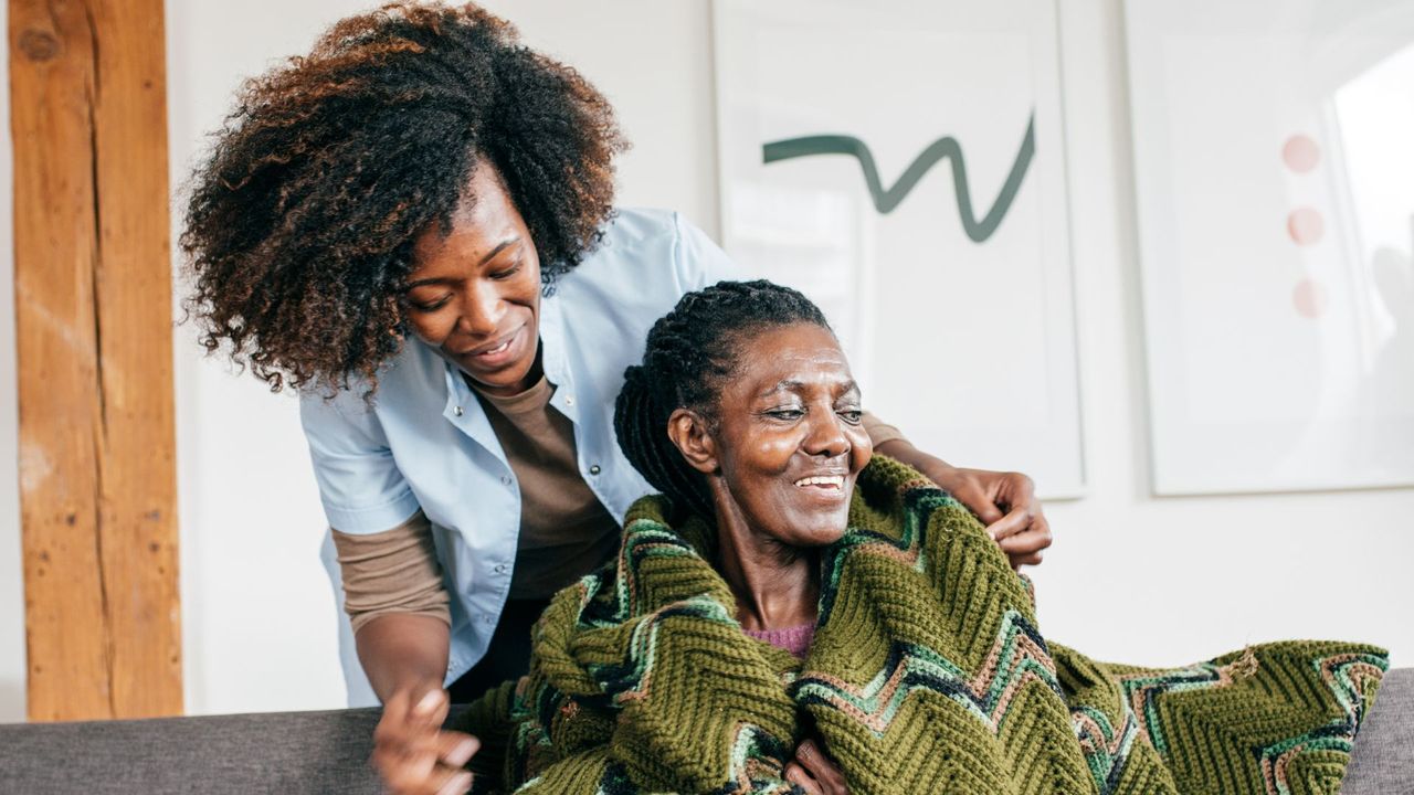 A caregiver from a home health care service helps a woman wrap a green patterned blanket around her shoulders in a cozy living room.