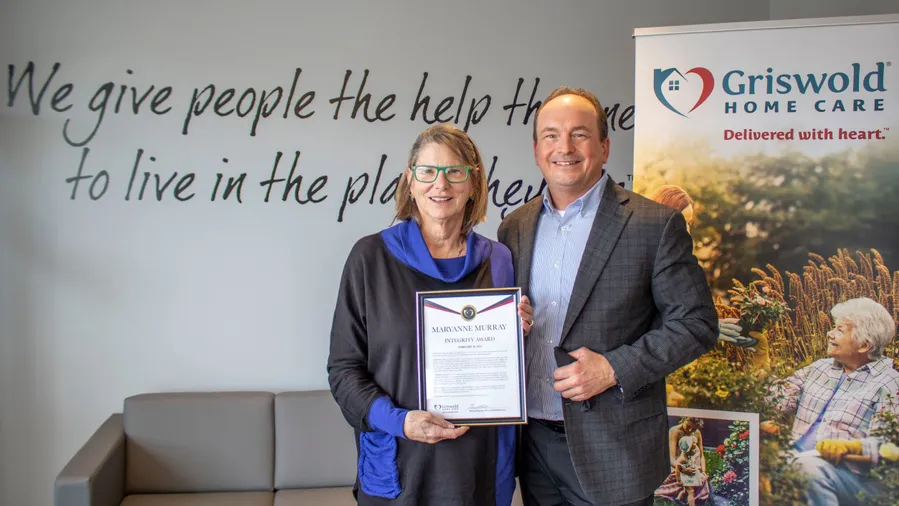 Two individuals stand indoors holding a certificate, smiling. The wall behind features text, and a banner with the Griswold Home Care logo is visible on the right, highlighting their exceptional caregiver services.