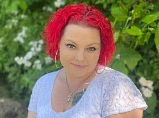 A person with bright red curly hair, wearing hoop earrings and a light-colored blouse, is posing outdoors in front of leafy greenery and white flowers, exuding a sense of well-being that reflects the importance of personal care services.