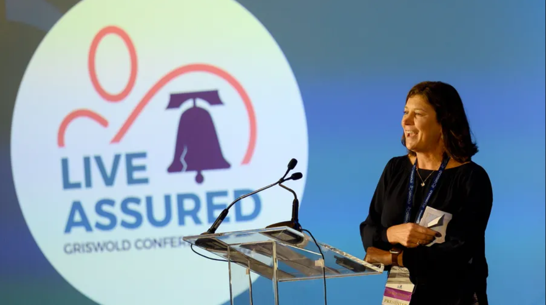 A person speaks at a podium on a stage with a "Live Assured Griswold Conference" sign in the background, highlighting the importance of caregiver services in enhancing quality of life.