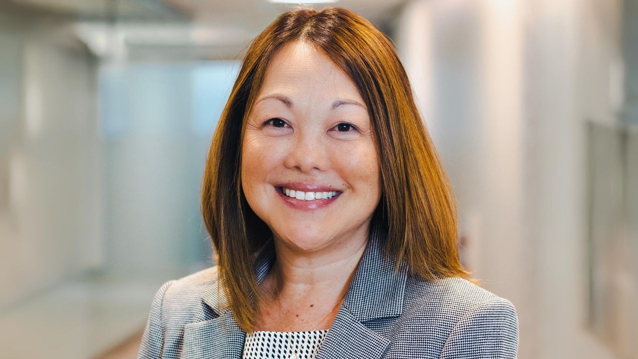 A person with shoulder-length brown hair and wearing a grey blazer and white patterned blouse is smiling in an indoor setting, reflecting the warmth often associated with home care.