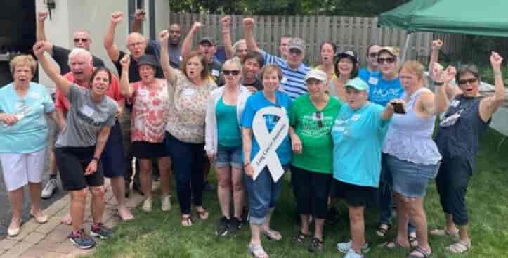 A group of people standing outdoors, many raising their fists in the air, some wearing teal clothing and ribbons, smiling at the camera. They appear to be at a gathering or event celebrating caregiver services.