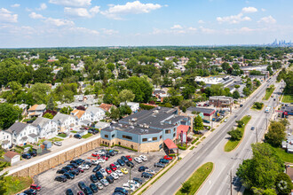 Aerial view of a suburban neighborhood with residential homes, a commercial building, and a parking lot near a busy street. The horizon shows a city skyline in the distance, with nearby facilities providing home health care services to the community.