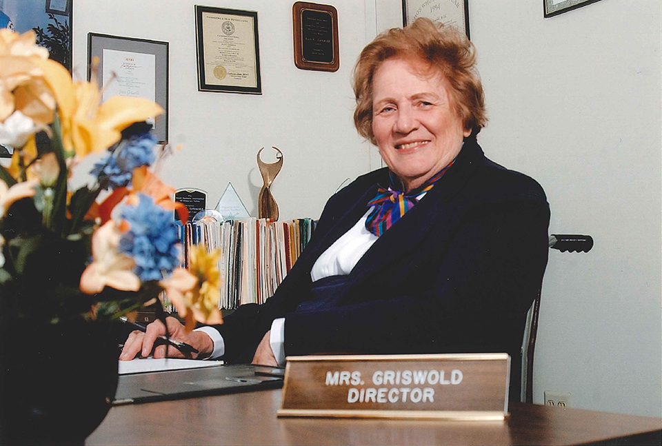 A woman sitting at a desk with a nameplate reading "Mrs. Griswold Director," flowers in the foreground, and awards on the wall behind her, overseeing elder home care services with dedication and compassion.