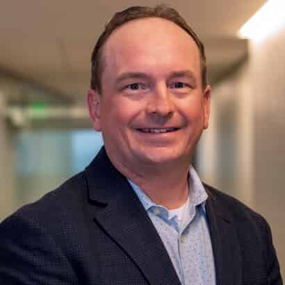 A man in a dark blazer and light blue shirt smiles while standing in a well-lit hallway, embodying the warmth and professionalism of our home health care service.