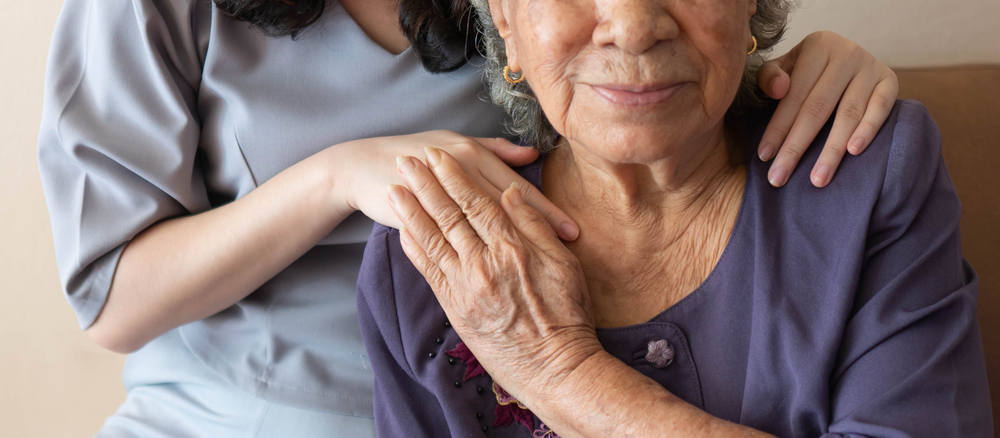 Close-up of a younger person gently placing their hands on the shoulders of an elderly woman who is sitting down, symbolizing compassionate caregiver services. Both are wearing purple and gray attire. The elderly woman has a calm expression.