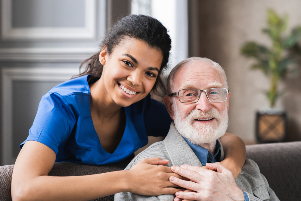 A young woman in a blue top smiles while standing behind and hugging an elderly man in glasses and a gray jacket, illustrating the warmth of caregiver services. They both look at the camera.