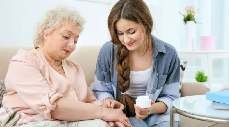 A young woman with a long braid applies lotion to an elderly woman's hand as they sit together on a couch in a brightly lit room, reflecting the gentle attentiveness typical of elder home care services.