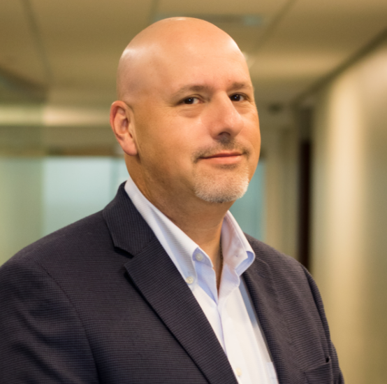 A bald man with a goatee, wearing a dark blazer and light-colored shirt, stands in a corridor, looking at the camera. His professional demeanor suggests expertise in personal care services.