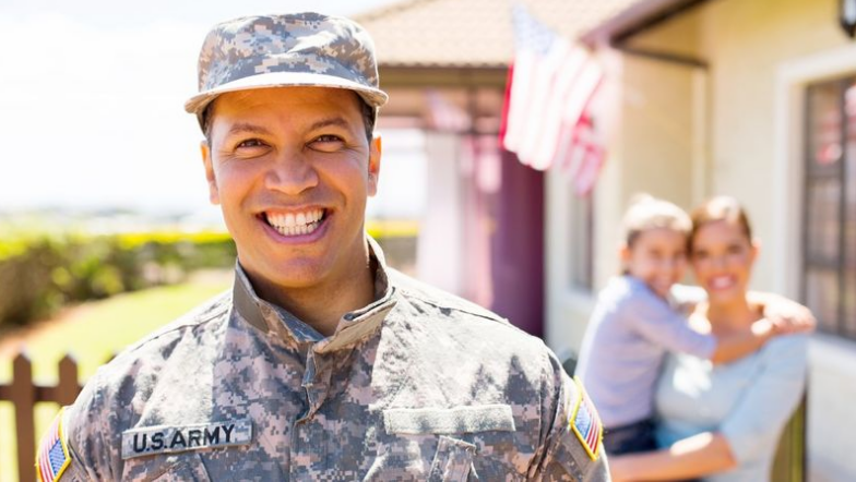 A U.S. Army soldier smiles in uniform outdoors with a blurred background of a woman holding a child and an American flag, symbolizing the deep commitment shared by those who serve and care for their communities, whether in uniform or through elder home care services.