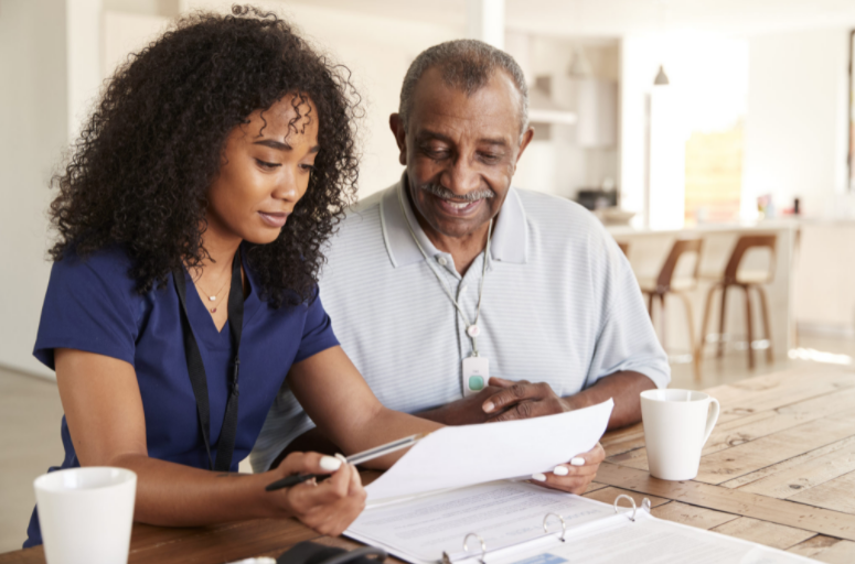 A woman and an older man sit at a table reviewing documents, likely related to elder home care services. They both wear lanyards and have coffee mugs in front of them. The background shows a kitchen area.