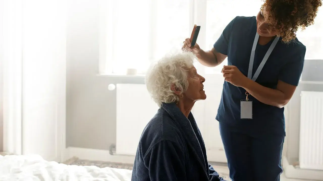 A caregiver combs the hair of an elderly person sitting on a bed