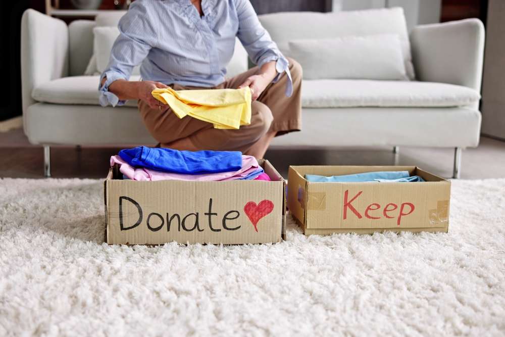 A person is sorting clothes, placing items into a cardboard box labeled "Donate" and another labeled "Keep." A white couch and a soft, white carpet are visible in the background.