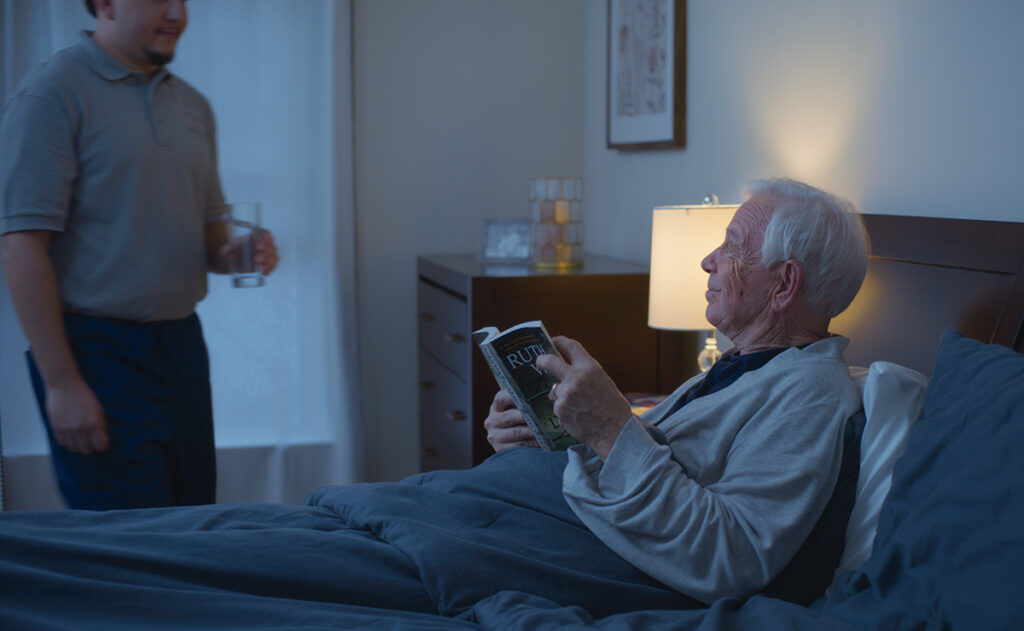 An elderly man with white hair reads a magazine in bed while a younger person, providing around-the-clock care, stands nearby holding a glass of water. A bedside lamp is lit, illuminating the room.